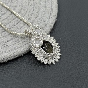 May include: A silver wire-wrapped pendant featuring a central oval black gemstone with gold-toned inclusions, accented by two small round white stones. The intricate metalwork creates a textured, radiating border, suspended from a silver chain.