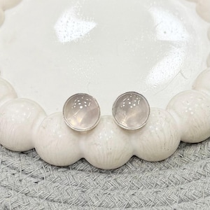 Op de afbeelding: Een paar ronde oorstekers met een lichtroze, doorschijnende steen gezet in een zilverkleurig metaal. De oorbellen worden gepresenteerd op een witte, decoratieve schaal met een parelrand, rustend op een grijs geweven oppervlak.
