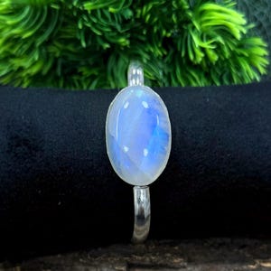 May include: A silver ring featuring a large, oval-shaped moonstone. The moonstone has a pearly, opalescent appearance with blue and white hues. The ring is set against a dark background with green foliage.