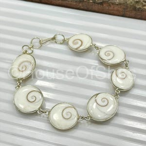 Op de afbeelding: Een zilveren armband met ovale en cirkelvormige witte schelpsegmenten met een spiraalvormig ontwerp. De segmenten zijn verbonden door zilveren schakels en een sluiting. De armband wordt getoond op een witte ondergrond.