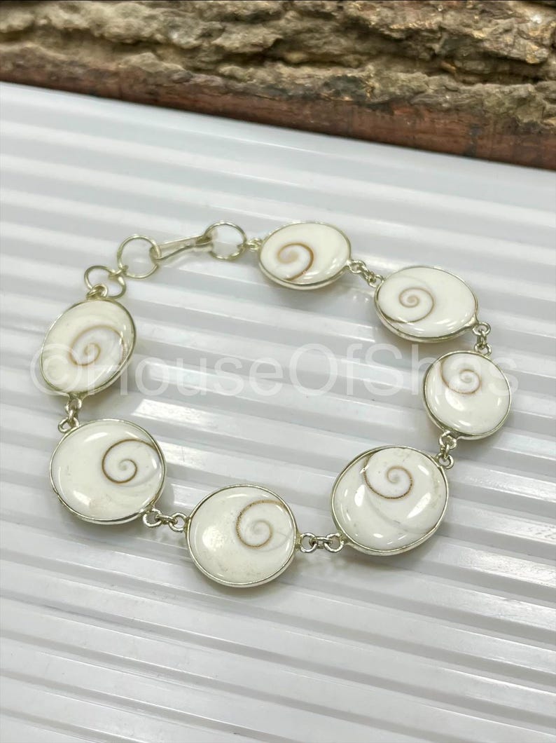 Op de afbeelding: Een zilveren armband met ovale en cirkelvormige witte schelpsegmenten met een spiraalvormig ontwerp. De segmenten zijn verbonden door zilveren schakels en een sluiting. De armband wordt getoond op een witte ondergrond.