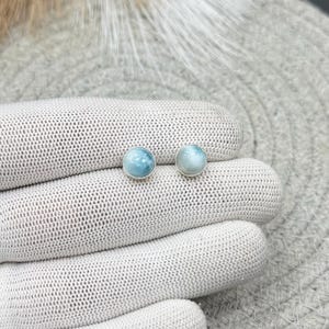 Könnte beinhalten: Ein Paar kleine, runde Ohrstecker mit polierten, hellblauen, marmorierten Steinen in schlichten, silberfarbenen Metallfassungen. Die Steine haben eine glatte, gewölbte Oberfläche mit subtilen Farb- und Texturvariationen, die ein sanftes Aussehen erzeugen.