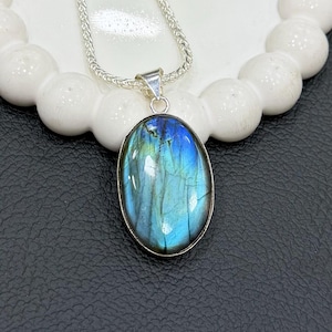 May include: A silver necklace featuring an oval labradorite pendant. The pendant has a silver bezel and displays iridescent blue and green hues. The necklace chain is a delicate silver snake chain. The pendant is displayed on a white, circular, beaded surface.