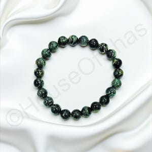 May include: A beaded bracelet featuring dark green and black speckled beads. The bracelet is displayed on a white satin surface, with two small, light brown stones in the background. The beads are approximately 0.5 inches in diameter.