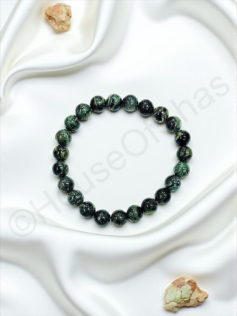 May include: A beaded bracelet featuring dark green and black speckled beads. The bracelet is displayed on a white satin surface, with two small, light brown stones in the background. The beads are approximately 0.5 inches in diameter.