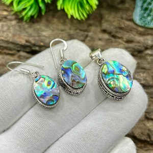 May include: Abalone shell jewelry set featuring a pair of earrings and a pendant. The oval-shaped abalone shells display iridescent blues, greens, and purples, set in ornate silver-toned metal. The earrings have hook closures.