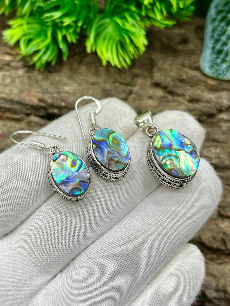 May include: Abalone shell jewelry set featuring a pair of earrings and a pendant. The oval-shaped abalone shells display iridescent blues, greens, and purples, set in ornate silver-toned metal. The earrings have hook closures.