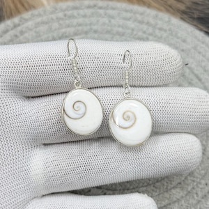 May include: A pair of silver earrings with white, circular pendants. Each pendant features a spiral design in the center. The earrings are displayed on a light gray surface.