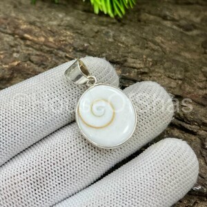 May include: A round, white pendant with a silver-colored bail. The pendant features a spiral design in the center, with a gold-colored outline. The pendant is set in a silver-colored metal.