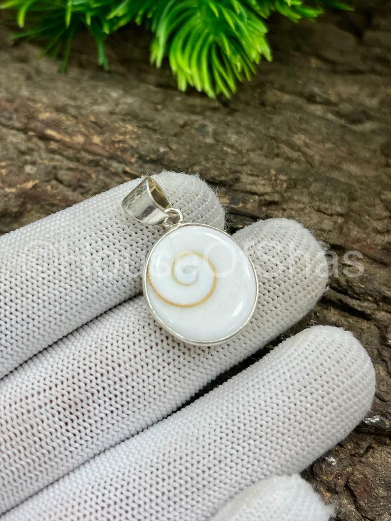 May include: A round, white pendant with a silver-colored bail. The pendant features a spiral design in the center, with a gold-colored outline. The pendant is set in a silver-colored metal.