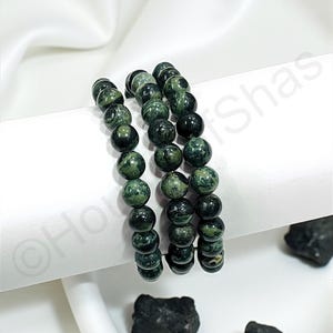 May include: Three beaded bracelets with dark green and black speckled stones. The bracelets are stacked on a white surface, with a white fabric backdrop. Several dark stones are scattered around the bracelets.
