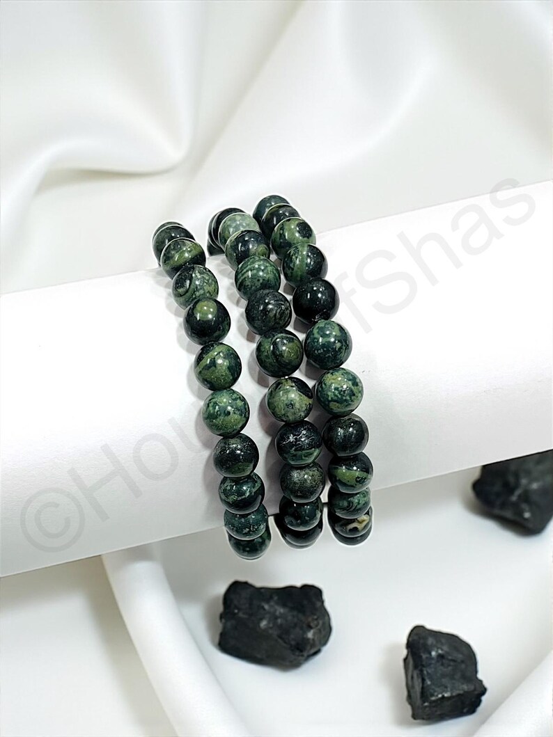 May include: Three beaded bracelets with dark green and black speckled stones. The bracelets are stacked on a white surface, with a white fabric backdrop. Several dark stones are scattered around the bracelets.