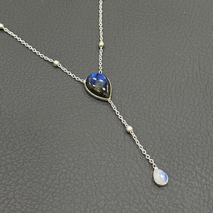 May include: A silver necklace with a teardrop-shaped pendant featuring a blue and black stone. The necklace has a delicate chain with small silver beads and a smaller teardrop-shaped stone at the end of a dangling chain.