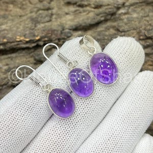 May include: A jewelry set featuring a pendant and earrings. The set includes a silver pendant and two earrings, each with a vibrant purple oval-shaped gemstone. The earrings have a coiled design above the gemstone.
