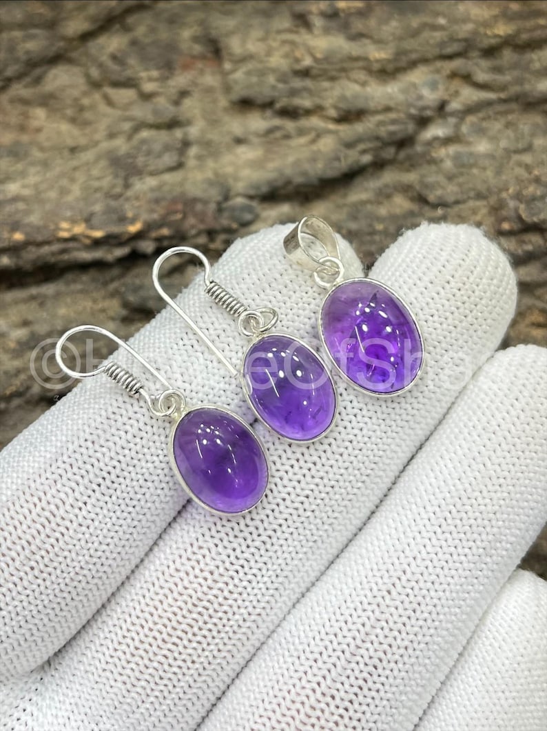May include: A jewelry set featuring a pendant and earrings. The set includes a silver pendant and two earrings, each with a vibrant purple oval-shaped gemstone. The earrings have a coiled design above the gemstone.