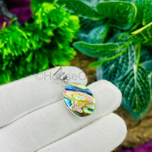 May include: Oval pendant with a silver bail, featuring a vibrant abalone shell inlay. The shell displays iridescent colors of blue, green, yellow, and pink. The pendant is held against a white glove, with green foliage in the background.