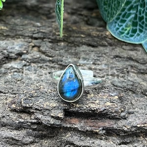 May include: A silver ring featuring a teardrop-shaped labradorite stone. The stone displays a vibrant blue and iridescent color. The ring is set against a textured, dark brown wooden surface, with green foliage in the background.