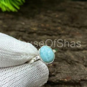 May include: A silver ring featuring a light blue oval gemstone. The ring is held by a gloved hand against a dark wood background. The gemstone is the focal point, showcasing a smooth, polished surface.
