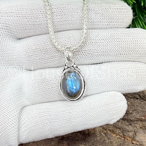 May include: A silver necklace with an oval pendant featuring a blue and gray gemstone. The pendant is set in a silver bezel and hangs from a delicate chain. The necklace is displayed on a white surface.