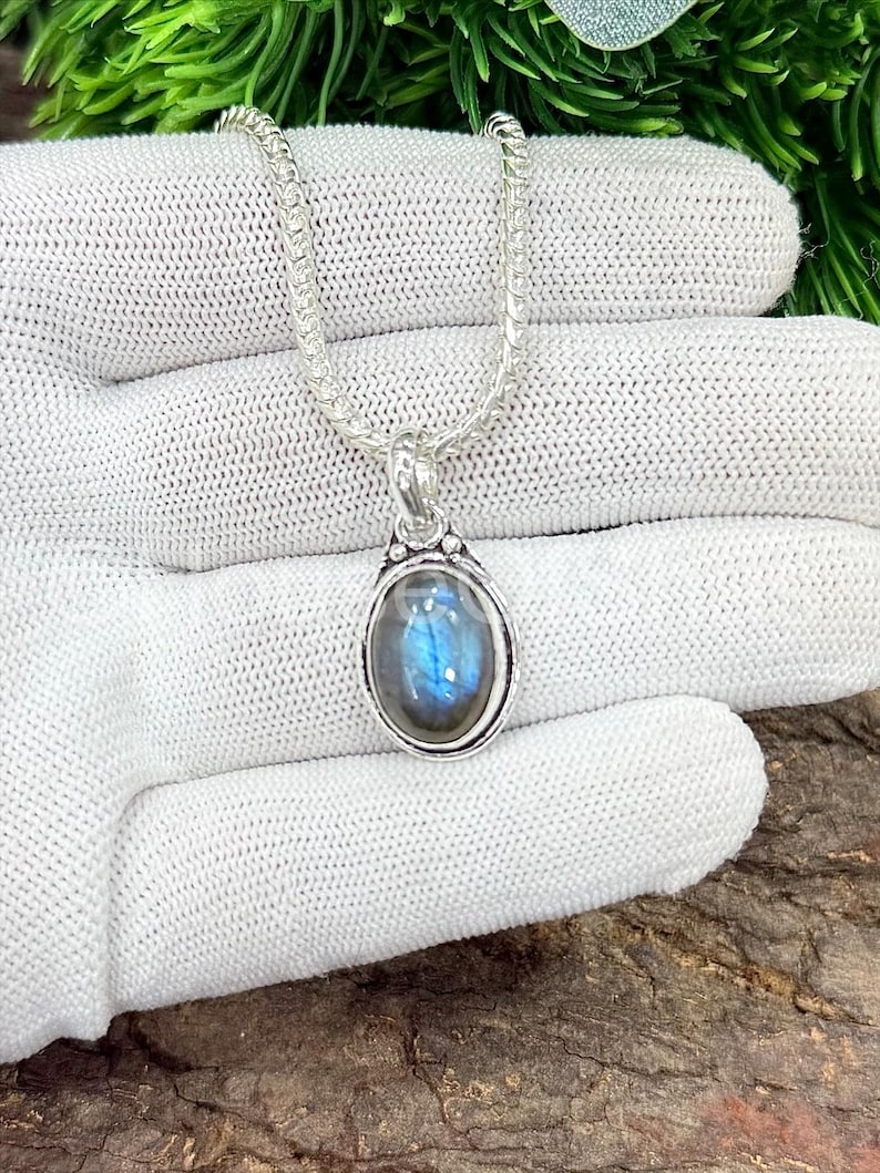 May include: A silver necklace with an oval pendant featuring a blue and gray gemstone. The pendant is set in a silver bezel and hangs from a delicate chain. The necklace is displayed on a white surface.
