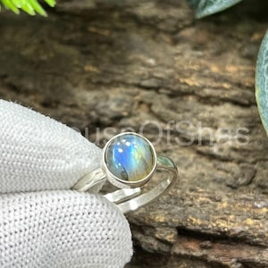 Puede incluir: Un anillo de plata con una piedra preciosa redonda, iridiscente, azul y amarilla. El anillo está sujeto por una mano enguantada. La piedra preciosa tiene un aspecto moteado. El anillo está sobre un fondo de madera natural.