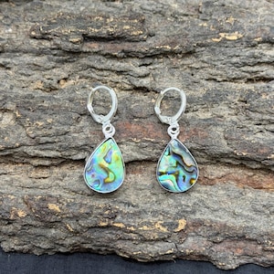 Może przedstawiać: Para kolczyków w kształcie łezki z iryzującymi muszlami z ucha morskiego osadzonymi w srebrnym metalu. Kolczyki zwisają z zawiasowych kółek. Muszle z ucha morskiego prezentują mieszankę kolorów zielonego, niebieskiego i złotego. Kolczyki są prezentowane na teksturowanej, brązowej powierzchni.