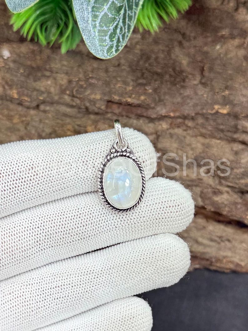 May include: Oval-shaped moonstone pendant with a silver-colored setting. The moonstone has a pearly, iridescent appearance. The pendant is suspended from a small loop at the top. The setting has a rope-like border.