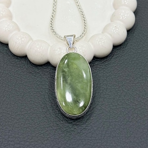 Op de afbeelding: Een zilveren ketting met een ovale, groene edelsteen hanger. De hanger is gezet in een zilveren rand en hangt aan een fijne zilveren ketting. De edelsteen heeft een gepolijste afwerking en de ketting wordt gepresenteerd op een wit, getextureerd oppervlak.