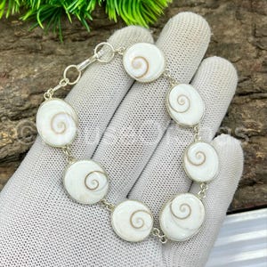 Op de afbeelding: Een zilveren armband met ovale hangers van witte en bruine spiraalvormige schelpen. De armband bestaat uit meerdere ovale hangers die met zilveren schakels zijn verbonden. De schelpen hebben een natuurlijk, wervelend patroon. De armband wordt getoond op een witte handschoen.