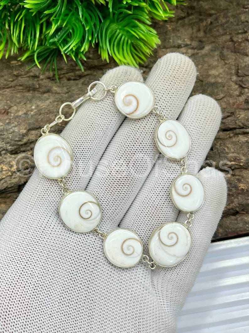 Op de afbeelding: Een zilveren armband met ovale hangers van witte en bruine spiraalvormige schelpen. De armband bestaat uit meerdere ovale hangers die met zilveren schakels zijn verbonden. De schelpen hebben een natuurlijk, wervelend patroon. De armband wordt getoond op een witte handschoen.