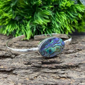 Pode incluir: Uma pulseira de punho prateada com uma pe&ccedil;a central oval de concha de abalone. A concha exibe iridesc&ecirc;ncias azuis, verdes e roxas. A pulseira est&aacute; sobre uma superf&iacute;cie de madeira escura texturizada, com folhagem verde ao fundo.