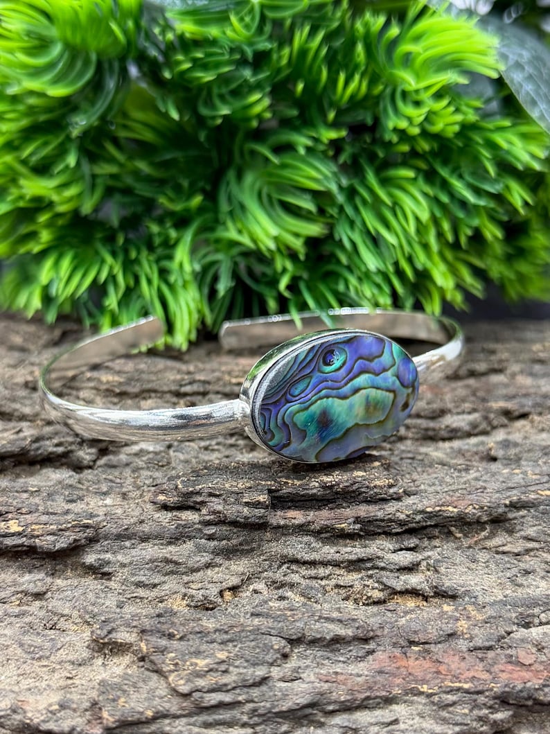 Pode incluir: Uma pulseira de punho prateada com uma pe&ccedil;a central oval de concha de abalone. A concha exibe iridesc&ecirc;ncias azuis, verdes e roxas. A pulseira est&aacute; sobre uma superf&iacute;cie de madeira escura texturizada, com folhagem verde ao fundo.