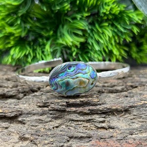 Pode incluir: Uma pulseira de prata com um centro oval de concha de abalone. A concha exibe azuis, verdes e roxos iridescentes. A pulseira est&aacute; sobre uma superf&iacute;cie de madeira texturizada e folhagem verde.