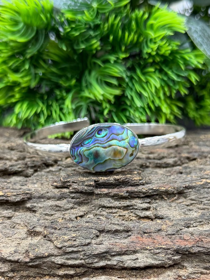 Pode incluir: Uma pulseira de prata com um centro oval de concha de abalone. A concha exibe azuis, verdes e roxos iridescentes. A pulseira est&aacute; sobre uma superf&iacute;cie de madeira texturizada e folhagem verde.