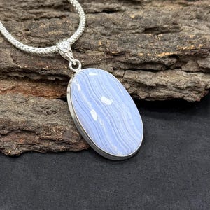 May include: A silver necklace with an oval pendant made of light blue lace agate. The stone displays white, wavy patterns. The necklace has a silver snake chain. The background is a dark, textured surface.