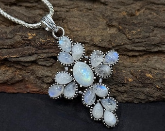Peut inclure: Un collier avec un pendentif en forme de croix argentée. La croix est ornée de plusieurs pierres de lune ovales et en forme de poire. Les pierres de lune sont d'un blanc laiteux avec une légère iridescence bleue. La croix est posée sur un fond de bois sombre.