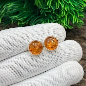 Puede incluir: Un par de pendientes de botón redondos de ámbar con engaste plateado. Los pendientes tienen un cálido tono naranja dorado y se muestran sobre una superficie de punto blanco, con follaje verde en el fondo. Joyas.