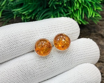 May include: A pair of round amber stud earrings with a silver setting. The earrings have a warm, golden-orange hue and are displayed on a white knitted surface, with green foliage in the background. The earrings are a jewelry item.