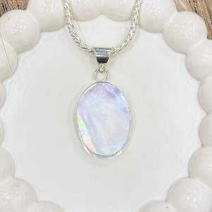 Op de afbeelding: Een zilveren ketting met een ovale, parelmoer hanger. De hanger heeft een glinsterend, iriserend oppervlak. De ketting is een gevlochten stijl. De ketting wordt gepresenteerd op een wit, geschulpte bord.