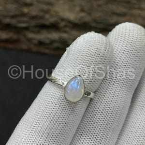 以下が含まれることがあります： 銀色の指輪。 涙の形をしたムーンストーンが特徴で、青と白の虹色の色合いを示しています。 リングは細いバンドです。 リングは白い布の上に表示されます。 宝石はシンプルなデザインにセットされています。
