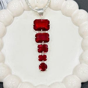 May include: A silver pendant necklace with a series of graduated, rectangular, and round, deep red gemstones. The gemstones are set in a silver metal setting and are connected by silver links. The necklace is displayed on a white, scalloped surface.