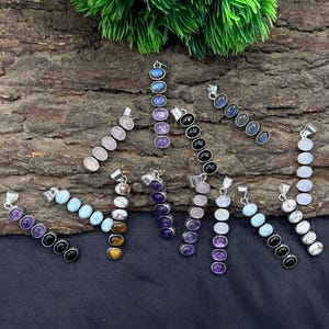 May include: A collection of gemstone pendants in various colors, including amethyst, labradorite, and rose quartz. Each pendant features a series of oval-shaped stones set in a silver-colored metal. The pendants are displayed on a textured wooden surface.