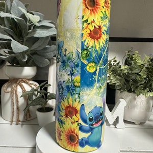 May include: A tall, cylindrical tumbler with a colourful design. The tumbler features sunflowers, blue floral elements, and a cartoon character. The colour palette includes yellow, blue, and white. The tumbler is likely made of stainless steel.