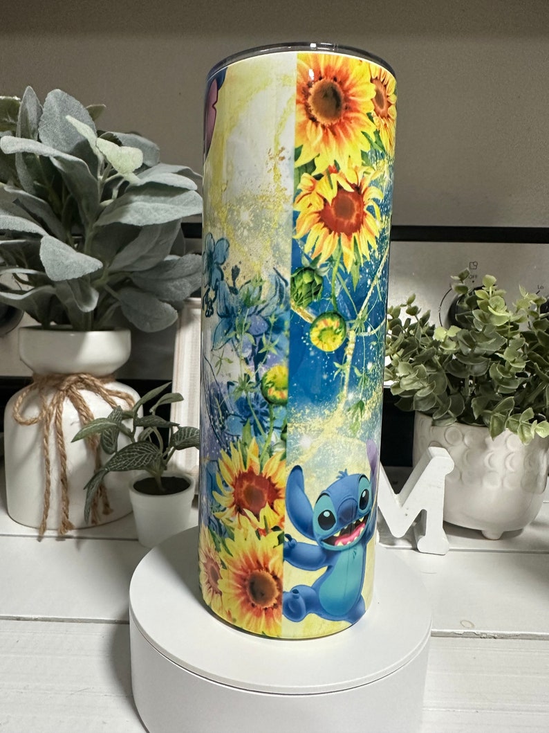 May include: A tall, cylindrical tumbler with a colourful design. The tumbler features sunflowers, blue floral elements, and a cartoon character. The colour palette includes yellow, blue, and white. The tumbler is likely made of stainless steel.