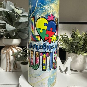 May include: A stainless steel tumbler with a lid, featuring the words "LOVE someone with AUTISM". The design includes colourful puzzle pieces, set against a blue and gold background. The tumbler is cylindrical in shape.