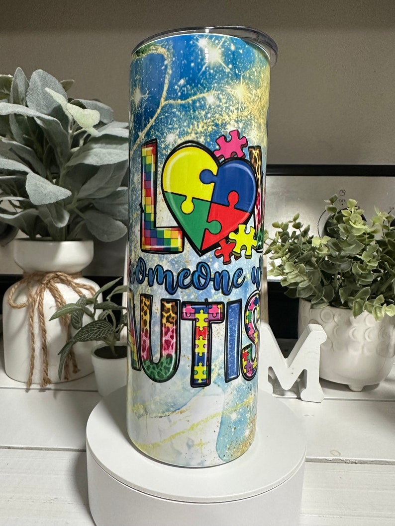 May include: A stainless steel tumbler with a lid, featuring the words "LOVE someone with AUTISM". The design includes colourful puzzle pieces, set against a blue and gold background. The tumbler is cylindrical in shape.