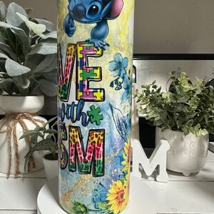 May include: A stainless steel tumbler with a vibrant design featuring the character Stitch. The tumbler displays the words "Love with Autism" alongside puzzle pieces, sunflowers, and blue floral patterns.