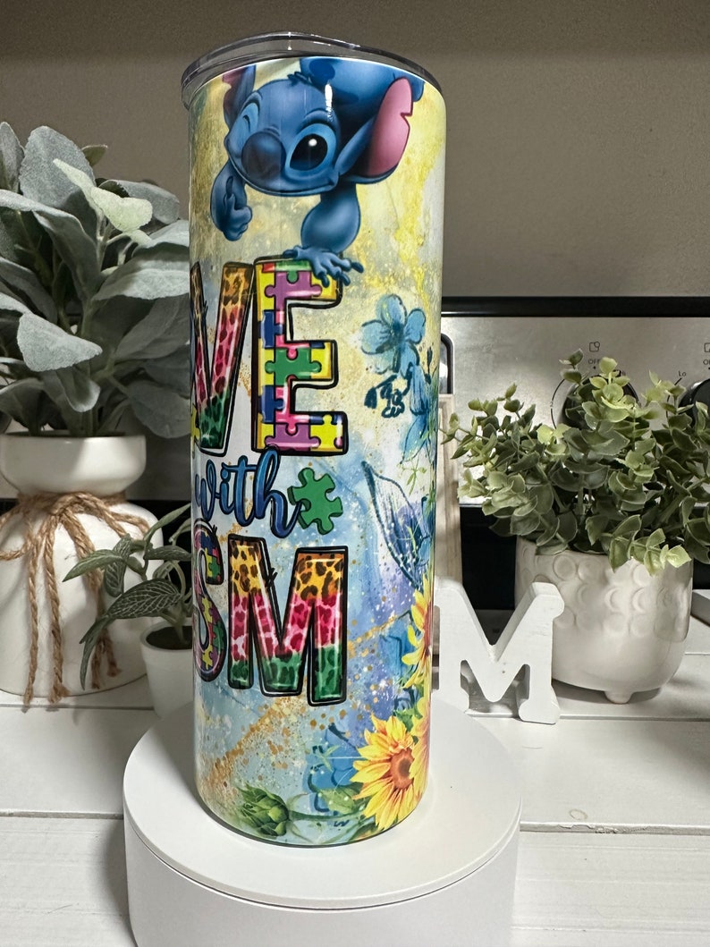May include: A stainless steel tumbler with a vibrant design featuring the character Stitch. The tumbler displays the words "Love with Autism" alongside puzzle pieces, sunflowers, and blue floral patterns.