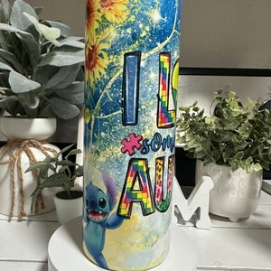 May include: A stainless steel tumbler with a vibrant design. The tumbler features sunflowers, a blue starry background, and the words "I LOVE SOME AU" with a puzzle piece and a cartoon character.