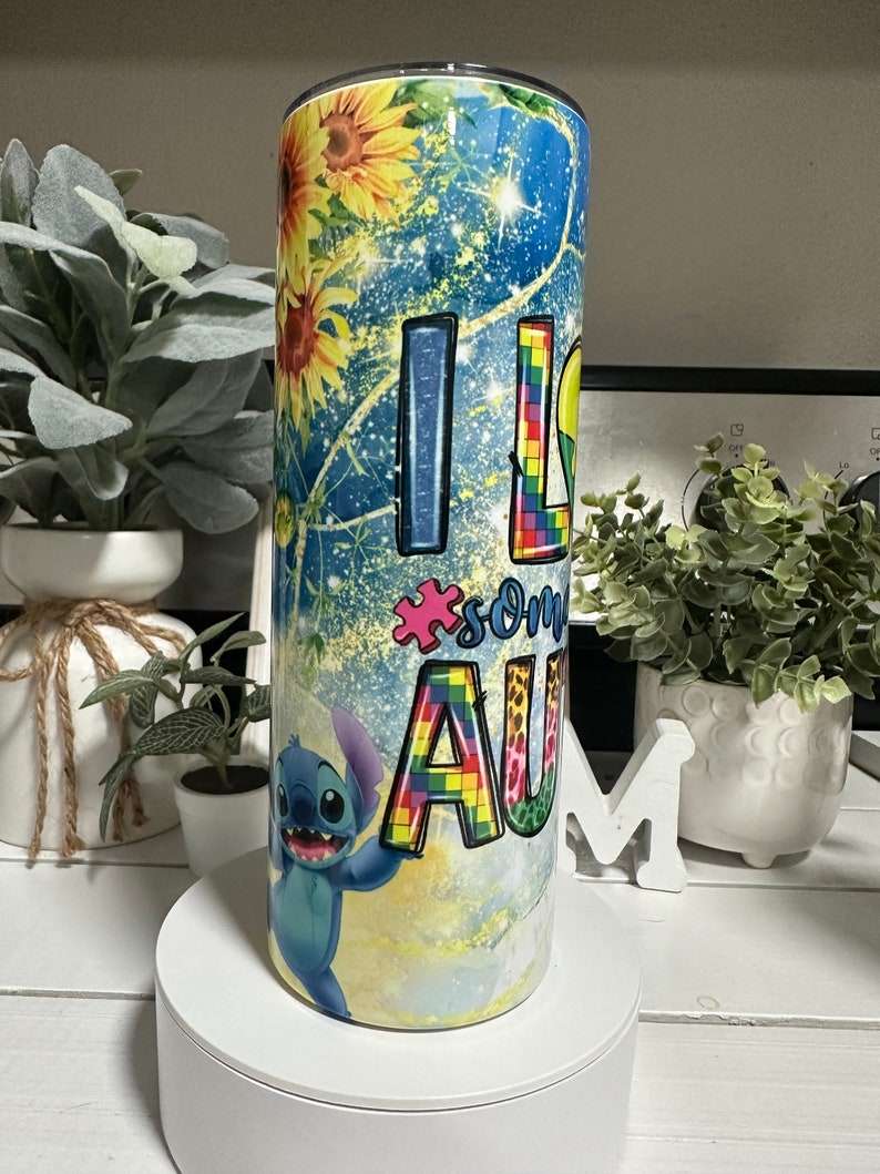 May include: A stainless steel tumbler with a vibrant design. The tumbler features sunflowers, a blue starry background, and the words "I LOVE SOME AU" with a puzzle piece and a cartoon character.